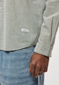 Svetlo zelena srajca z gumbi, teksturiranega zaključka, s etiketo na spodnjem delu spredaj in modnim detajlom na manšeti. Oblečena z modrim jeansa.