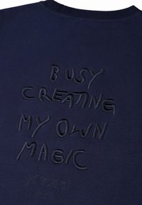 Felpa navy con testo ricamato che recita "IMPEGNATO A CREARE LA MIA MAGIA" con un accento decorativo sottostante. Tessuto morbido e testurizzato.