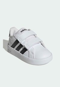 Bijela tenisica s crnim uzorkom od tri pruge, izrađena od sintetičke kože. Učvršćena s dva Velcro remena, gumenim potplatom. Zaobljen oblik vrha.