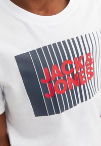 Balta kokvilnas T-kreklā ir liels grafiskais attēls ar "JACK & JONES" treknām sarkaniem burtiem un vertikālām jūras zilām svītrām fona apakšā.