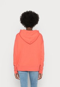 Persona con afro che indossa un hoodie corallo, di spalle, abbinato a jeans azzurri su uno sfondo bianco semplice.