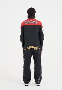 Svart och röd motorcykeljacka med vita ärmar; har "Ed Hardy"-logotyp i kursiv. Svarta byxor med broderade mönster och logotyp.
