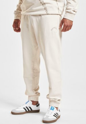 Beige Baumwoll-Sweatpants mit elastischen Bündchen, die ein dezentes Logo auf dem Oberschenkel tragen. Kombiniert mit weißen und schwarzen Sneakers.