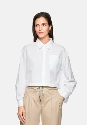 Frau trägt ein weißes, kurzes Hemd mit Knöpfen und beige Hose mit Kordelzug, steht mit einer Hand in der Tasche vor einem einfarbigen Hintergrund.