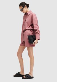 Conjunto de camisa y pantalones cortos de color rosa empolvado y de corte amplio, fabricado con una tela suave. Combinado con un bolso tipo clutch negro texturizado y sandalias planas negras.