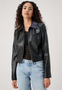 Giacca cropped in finta pelle nera con colletto a punta, chiusura con zip e taschino sul petto, abbinata a jeans di denim azzurro chiaro.