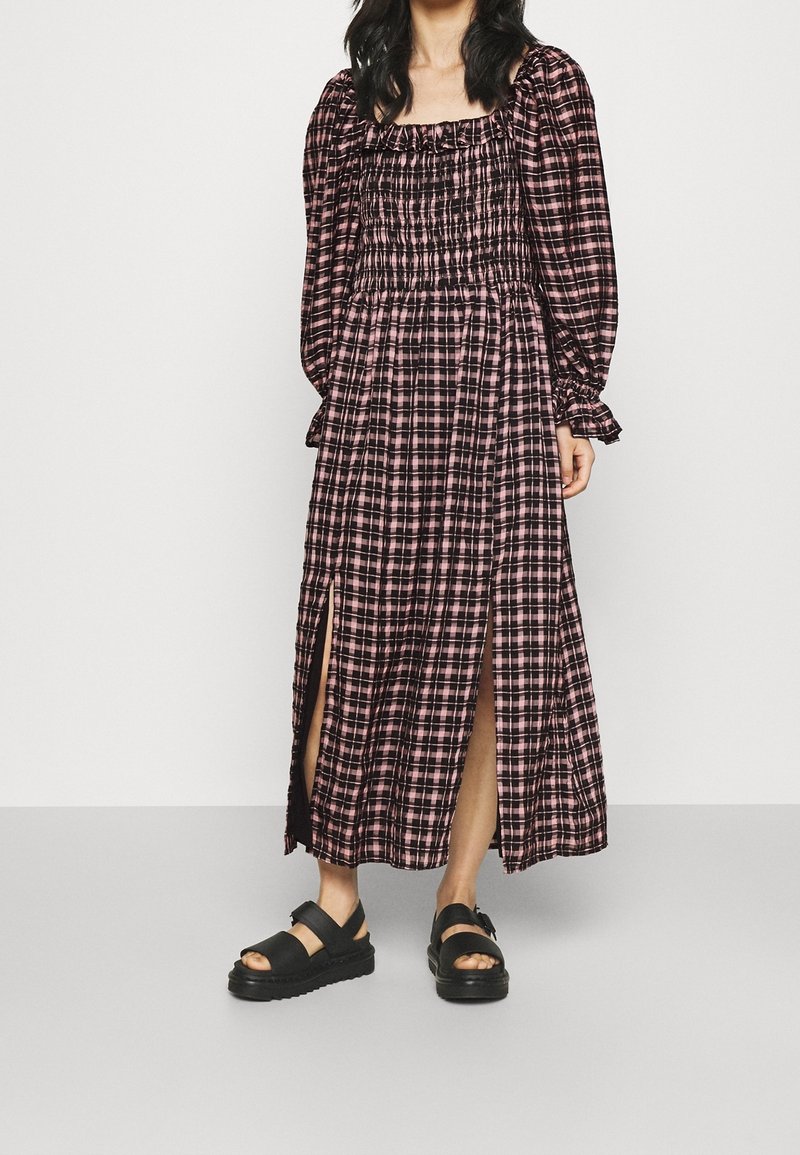Femme portant une longue robe à carreaux rose et noire avec des fentes et des sandales compensées noires, debout sur un fond blanc uni.