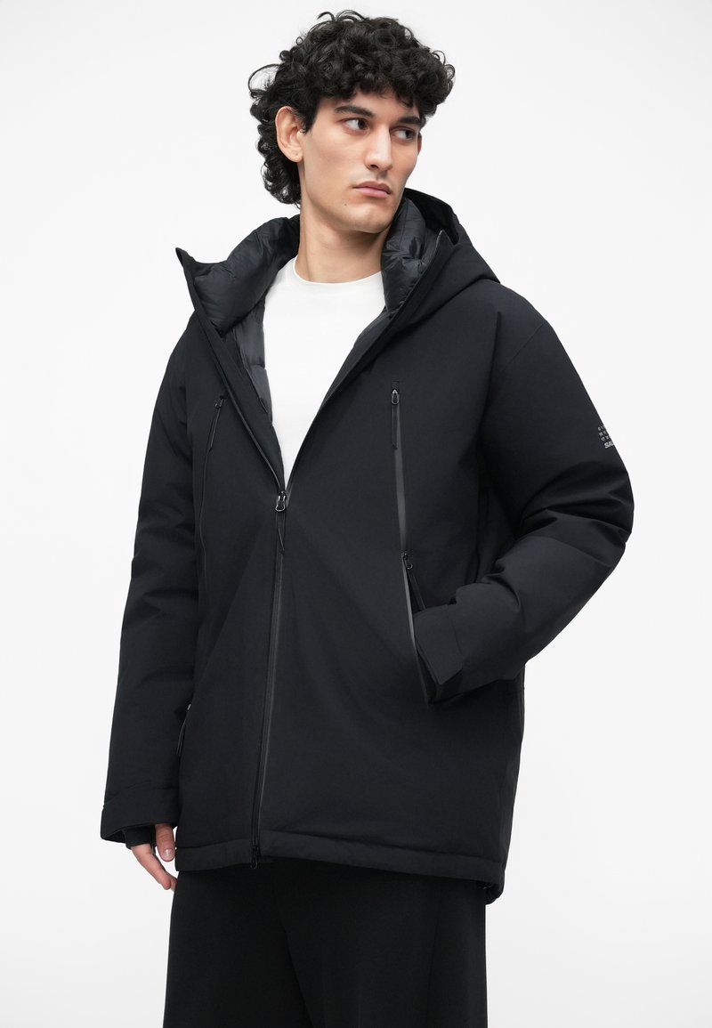 Sort isoleret jakke med hætte, der har lynlåslommer og en glat overflade. Enkel design med en afslappet pasform og minimale detaljer.