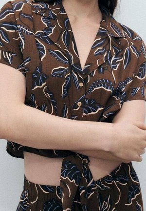 Femme portant une chemise marron à manches courtes et une jupe assortie avec des motifs de feuilles bleues et beiges, bras croisés sur le ventre.