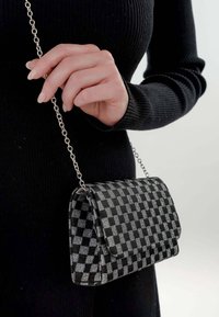 Bolso a cuadros negro y plateado con textura suave, cierre de solapa y strap de cadena metálica sostenido por una mano.
