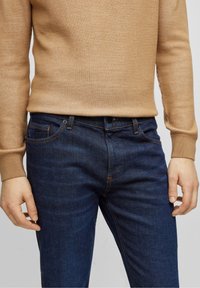 Béžový žebrovaný svetr a tmavě modré džíny, s viditelnými švovými detaily a střih přiléhavý, ukazující texturu a hladkou látku.