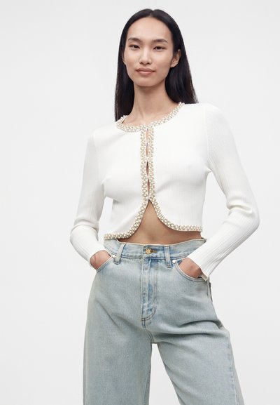 Femme portant un cardigan blanc court avec bordure en perles et un jean taille haute bleu clair, debout les mains dans les poches devant un fond uni.