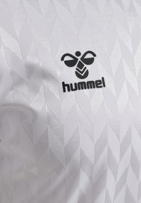 Maglietta sportiva bianca con un motivo a diamante testurizzato. Presenta un logo "hummel" nero e una grafica stilizzata di un'ape. Realizzata in materiale sintetico.