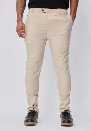 Homme portant un pantalon beige slim-fit et des chaussures noires, debout les mains dans les poches devant un fond uni.