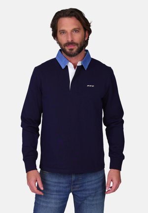 Homme barbu portant un maillot de rugby bleu marine à manches longues avec un col bleu et un petit logo, associé à un jean bleu, debout devant un fond blanc.