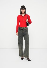 Polo à manches longues rouge avec trois boutons, associé à un jean en denim noir délavé à coupe ample et des chaussures à talons noires. Petit sac à main noir.
