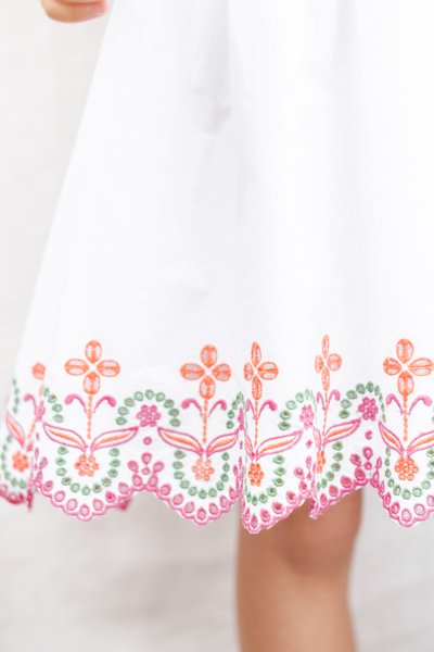 Λευκό φόρεμα από ύφασμα με πολύχρωμο κεντητό λουλουδάτο σχέδιο σε πορτοκαλί, ροζ και πράσινο κατά μήκος της πλαϊνής κυματιστής γραμμής. Λείος υφής, μήκος μέχρι το γόνατο.