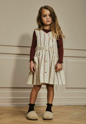 Ragazzina con i capelli lunghi che indossa un vestito a righe crema e marrone sopra una maglietta a maniche lunghe bordeaux e pantofole pelose, in piedi dentro casa.