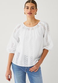 White embroidered blouse with a gathered neckline, balloon sleeves, and cut-out patterns along the front and sleeves. Paired with blue jeans.