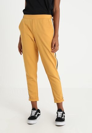 Person wearing mustard yellow pants with black and white side stripes, black top, and black-and-white sneakers, standing on white floor.