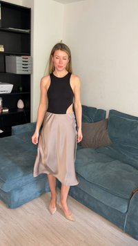 Femme portant un haut noir sans manches et une jupe en satin beige, debout pieds nus sur un sol en bois clair dans un salon avec un canapé bleu et des étagères noires.