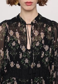 Blouse noire à fleurs en tissu transparent, dotée d'un design froncé, d'accents en dentelle, d'un col à nouer, et de motifs floraux rose clair et vert.