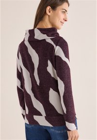 Sweter z cowl-neck w głębokim burgundzie, z wyrazistymi białymi abstrakcyjnymi paskami. Miękka faktura z luźnym krojem i ściągaczem.