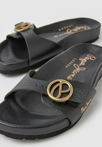 Sandali in pelle nera con una fascia larga e una fibbia a forma di simbolo della pace in oro. La soletta è strutturata e la suola è leggermente rialzata.