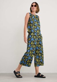 Jumpsuit bez rukávů s květinovým vzorem v černé, zelené a modré barvě. Volný střih s širokými zkrácenými kalhotami. Nosit s černými plochými sandály a slunečními brýlemi.
