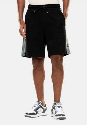 Hombre vistiendo pantalones cortos negros con paneles laterales de rayas verticales y zapatillas EA7 blancas y negras, de pie contra un fondo liso.