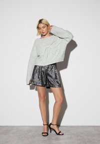 Grauer gestrickter Pullover mit einer Schleifendetail, kombiniert mit glänzenden schwarzen Shorts; das Model trägt schwarze Riemchenabsätze. Schlichter Hintergrund.