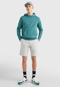 Felpa con cappuccio verde acqua con tasca frontale e logo in rilievo, abbinata a pantaloni corti chiari. Modello che indossa scarpe da ginnastica bianche e verde acqua con calzini a righe.