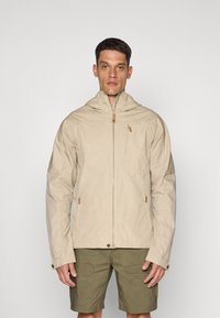 Veste légère beige avec une capuche, fermeture éclair, poches latérales et détails en bois. Portée sur un short vert olive.