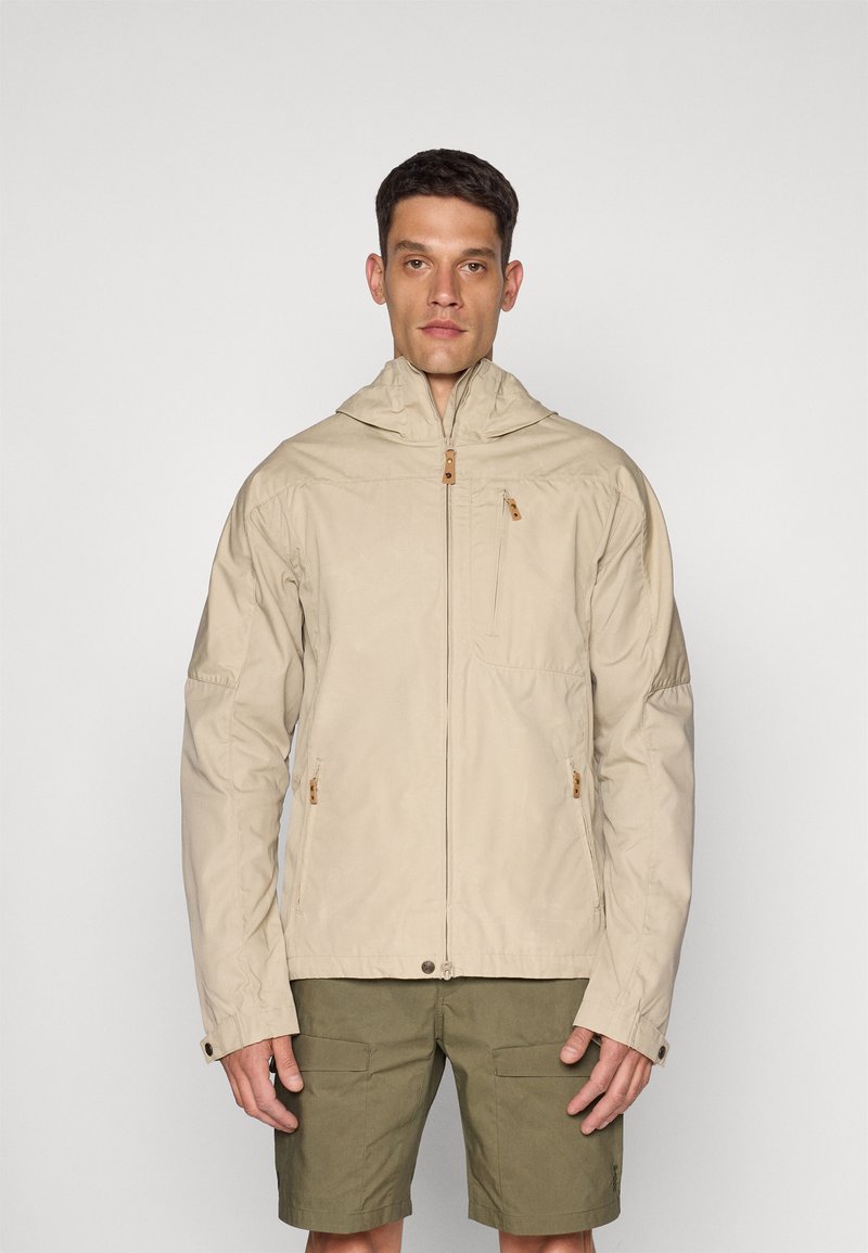 Veste légère beige avec une capuche, fermeture éclair, poches latérales et détails en bois. Portée sur un short vert olive.