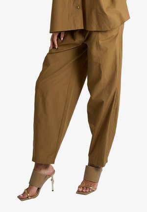 Donna con pantaloni marroni larghi e sandali beige con tacco alto e cinturini dorati, in piedi con una mano appoggiata sulla coscia.