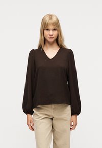 Ξανθιά γυναίκα που φοράει μπλούζα μακρυμάνικη καφέ σκούρου χρώματος με V-λαιμόκοψη και φουσκωτά μανίκια, καθώς και μπεζ παντελόνια, στέκεται μπροστά σε απλό φόντο.