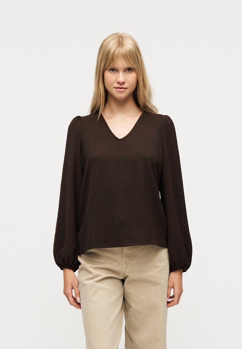 Ξανθιά γυναίκα που φοράει μπλούζα μακρυμάνικη καφέ σκούρου χρώματος με V-λαιμόκοψη και φουσκωτά μανίκια, καθώς και μπεζ παντελόνια, στέκεται μπροστά σε απλό φόντο.