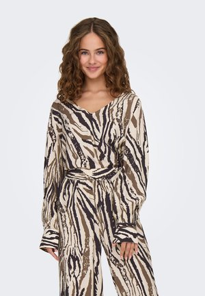 Ung kvinde med krøllet hår iført matchende beige, brun og sort zebra-stribet langærmet top og bukser, smilende til kameraet.