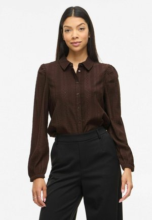 Ženska z dolgimi temnimi lasmi, oblečena v temno rjavo teksturirano bluzo z gumbi in nabranimi rokavi ter črne hlače z visokim pasom.