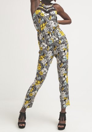 Femme portant une combinaison sans manches à fleurs jaunes, blanches et bleues, posant avec une main sur la hanche et des talons noirs à franges.