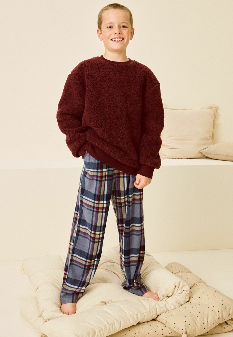 Maroon fleece-huppari ja siniset ruudulliset pyjamahousut lapsella, joka seisoo vaalealla tyynyllä. Yksinkertainen tausta tyynyineen.