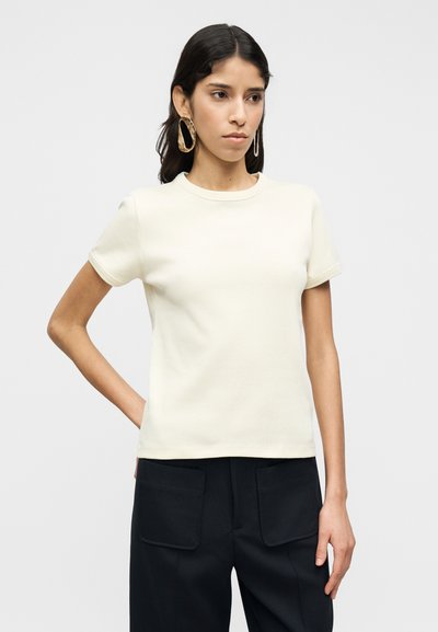 Μπλούζα κοντομάνικη, από βαμβάκι χρώματος κρέμα, με στρογγυλή λαιμόκοψη. Το ύφασμα είναι λείο με ελαφρώς εφαρμοστή σχεδίαση. Φορεμένη με σκούρο παντελόνι.
