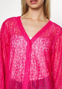 Cardigan in pizzo rosa con scollatura a V profonda, maniche lunghe e chiusura a bottoni. Il tessuto è trasparente con una texture uniforme.