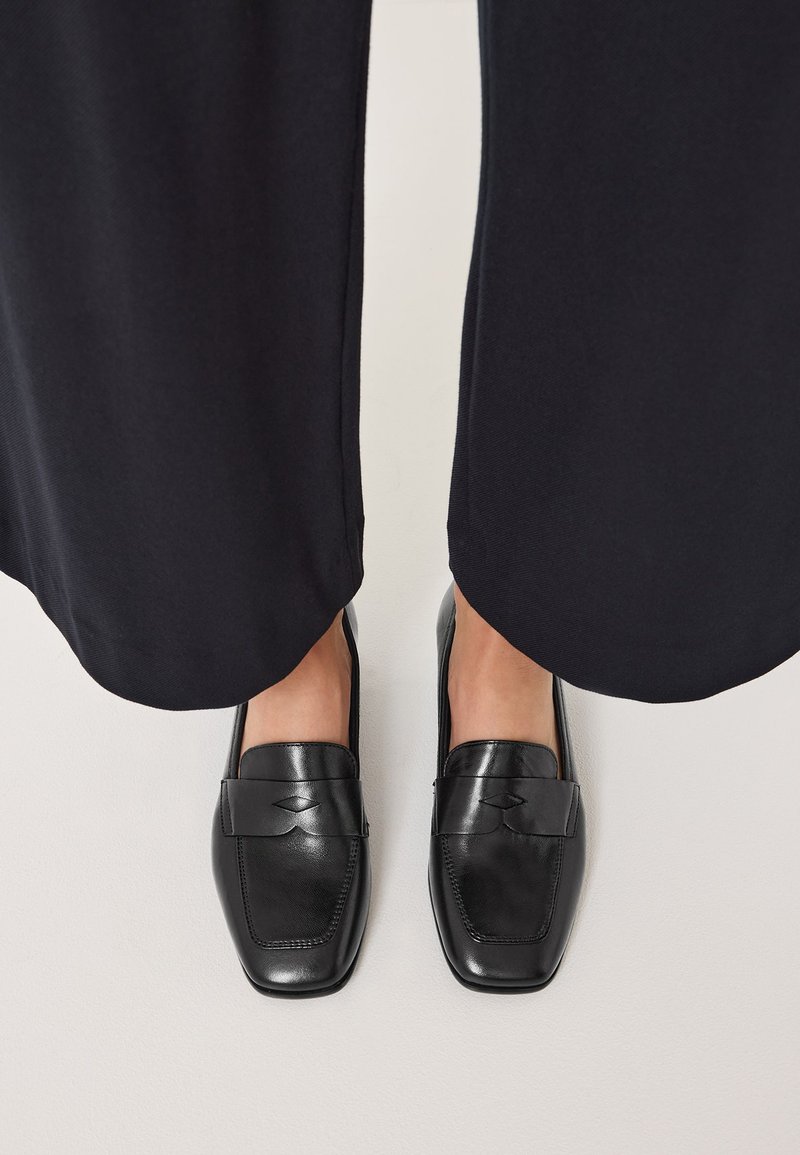 Sorte læder loafers med rektangulær form, glat texture og et dekorativt bånd over fronten. Båret med mørke bukser.