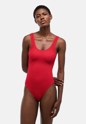 Femme aux cheveux courts portant un body rouge texturé sans manches, debout avec un bras plié derrière le dos.