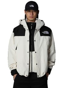 White and black windbreaker jacket with a hood, zippered front, and snap button closure. Features logo on the chest and textured fabric.