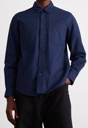 Mann trägt ein dunkelblaues Hemd mit Knöpfen und zwei Brusttaschen sowie eine schwarze Hose, eine Hand in der Tasche, vor einem schlichten Hintergrund.