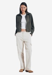 Zielona zamszowa krótka kurtka z dwoma przednimi kieszeniami, zestawiona z beżowymi spodniami cargo o wysokim stanie i brązowymi butami na obcasie.