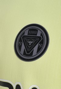 Un patch circulaire noir sur un tissu texturé jaune vif, avec un emblème triangulaire et le nombre "XXIII" en haut.
