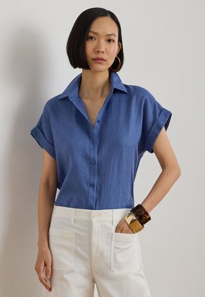 Cămașă albastră cu mâneci scurte, cu nasturi pe față, confecționată dintr-un material moale, asortată cu pantaloni albi și brățări groase. Textură netedă, croială casual.